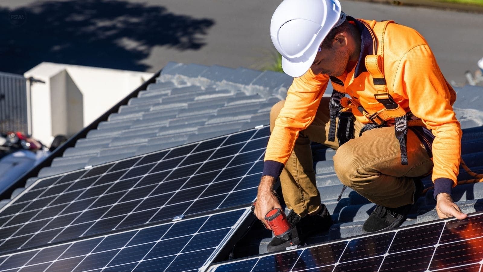Installer wearing a hard hat and safety harness fastening a rooftop solar panel.