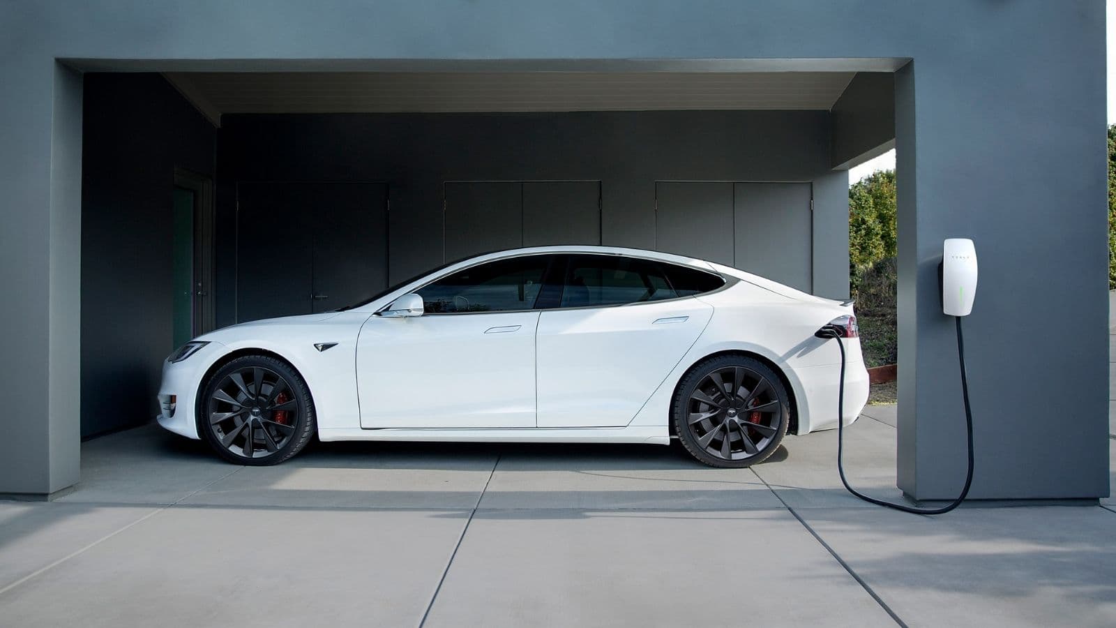White Tesla plugged into a wall-mounted home charger beside a garage.