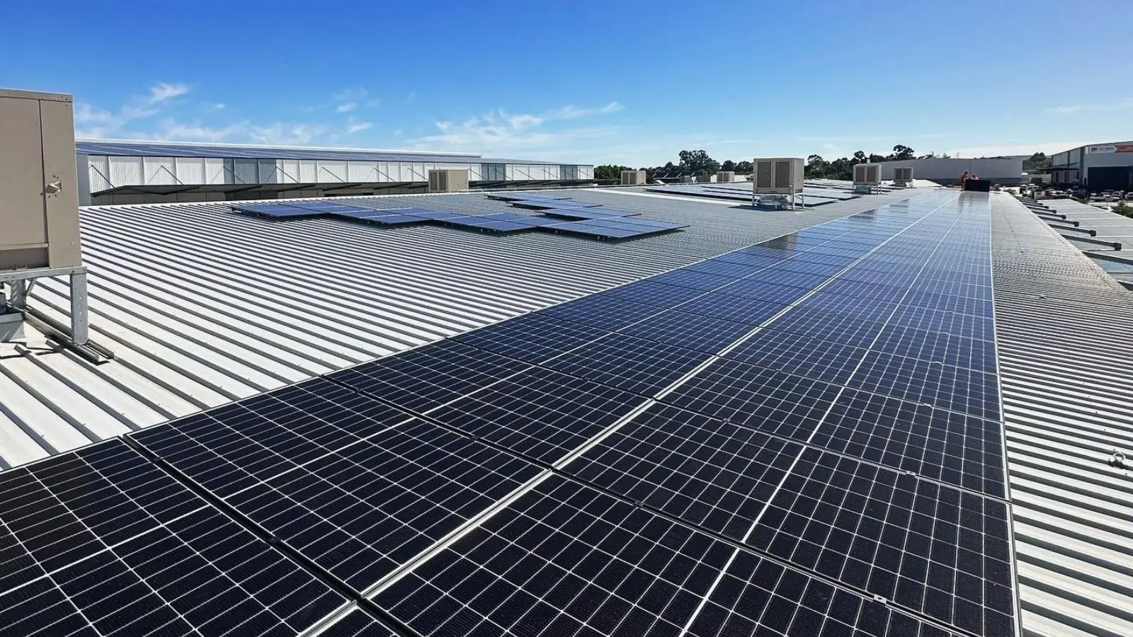 Solar panels across a corrugated commercial rooftop, with rooftop air-conditioning units under a blue sky.