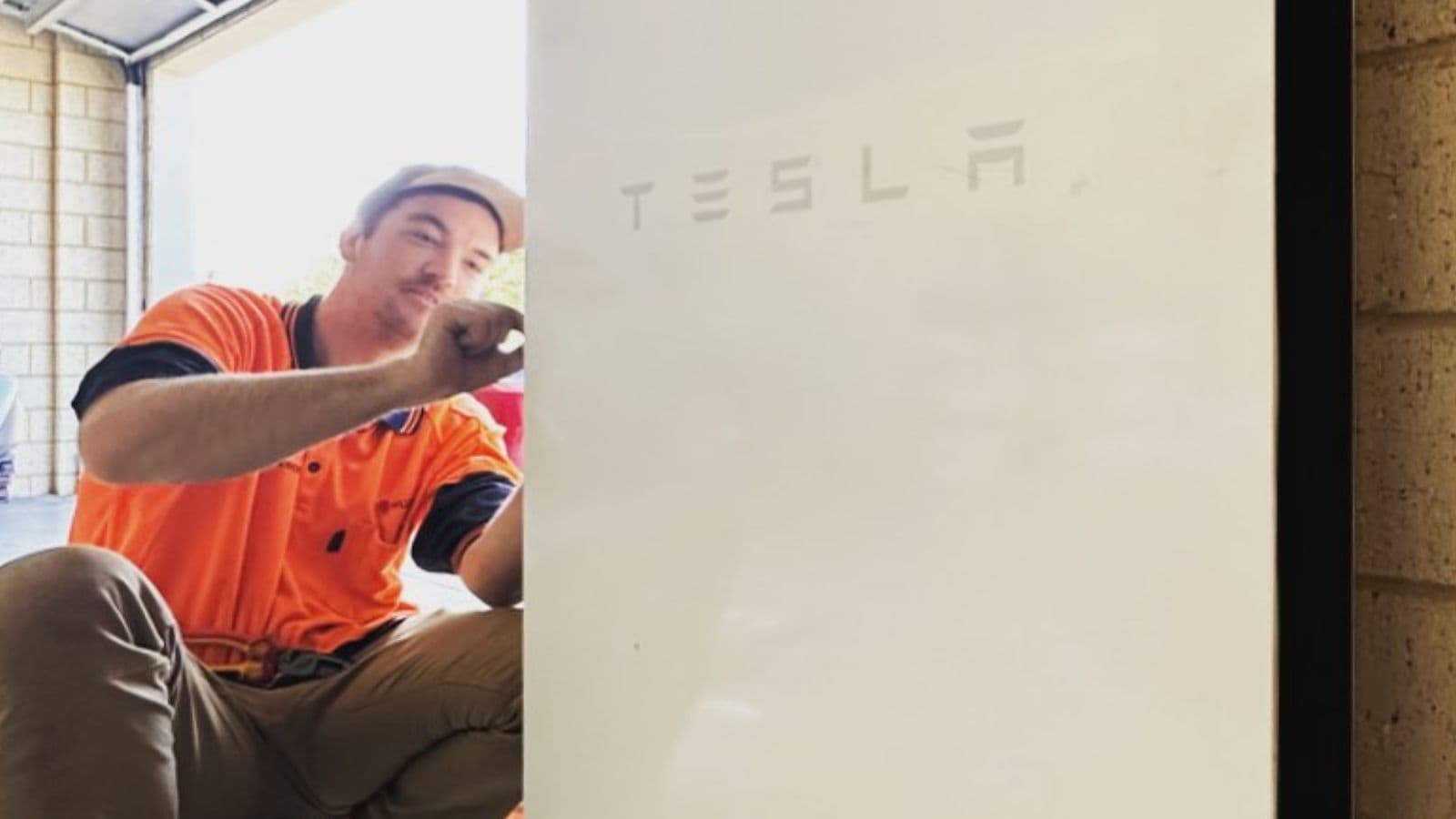 Installer in an orange work shirt beside a white Tesla Powerwall battery in a garage.