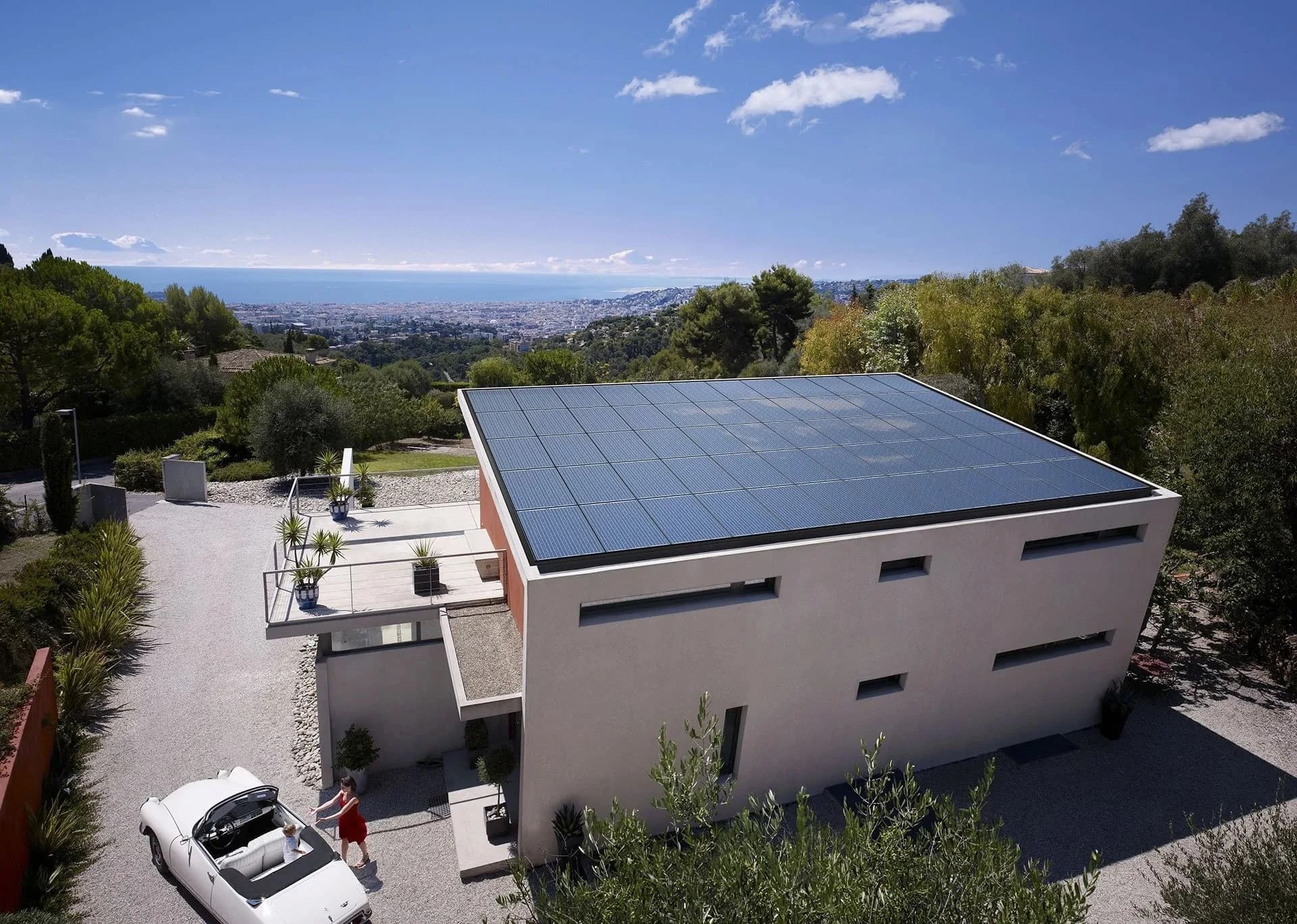Whole roof top covered in black solar panels over looking a beach in the distance. Young mother and child in a classic convertible car at the left of the house.