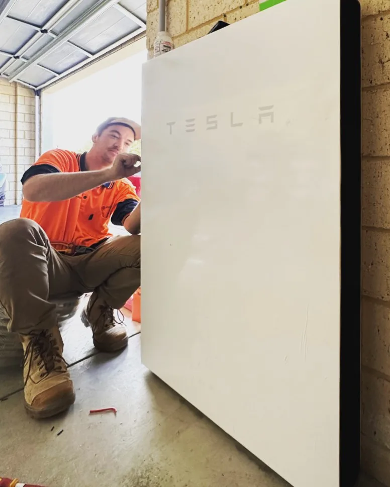 A PSW installation technician working beside a wall-mounted Tesla Powerwall at a brick home